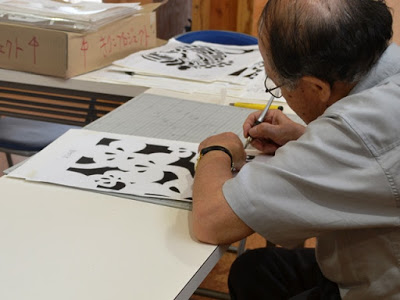 年配の男性がカッターナイフで黒と白の切り絵の紙を丁寧に切り抜いている作業中の写真