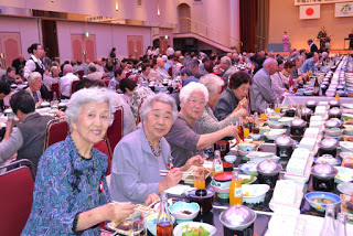 手前の席で数人の高齢の女性たちが笑顔でカメラに向かいながら食事を楽しんでおり、背景には多くの人々が座っている写真