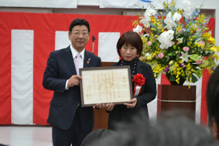 紅白幕が飾られ、大きな花の飾りが置かれている前で、佐藤町長と左胸に赤い胸章リボンとつけた女性が並び、一緒に額縁に入った感謝状を持っている表彰式の写真