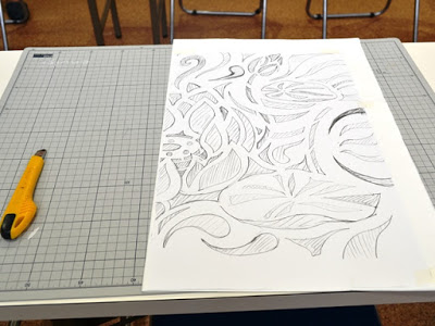 カッターマットの上に花や葉のような模様が鉛筆で描かれた白い紙が置かれていて、左には黄色いカッターが置かれている下絵段階の作品の写真