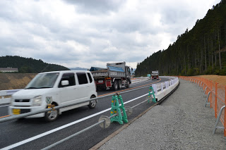曇り空の下、右側には森林、左側には開けた土地が広がる片側一車線の道路を、左側に白い軽自動車、右側に2台のダンプカーが連なって走る対面通行の道路が写されており、道の両脇には緑色の工事用バリケードやオレンジ色のフェンスが設置された工事中の道路の写真