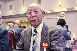 グレーのチェック柄スーツに青いネクタイを着用し、胸に赤白のリボンをつけた高齢の男性が正面を向いて落ち着いた表情で座っている敬老会会場内の写真