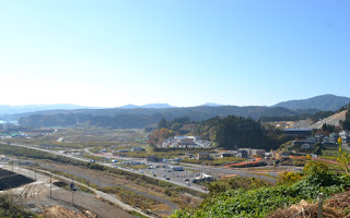 青空の下に山々が広がり、その手前の谷には道路や建物が点在していて、複数の車が走っており、緑の木々がある町の様子を高台から撮影した写真