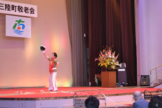 ステージ中央で白いズボンと赤い衣装を着た女性が帽子を片手に掲げてパフォーマンスを行っている写真