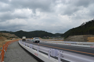 曇り空の下、白いトラックや乗用車が走る対面通行の新設道路が写されており、両側にはガードレールが設置され、左側にはオレンジ色の安全フェンスが並び、背景には木々に覆われた丘陵地が広がる道路風景の写真