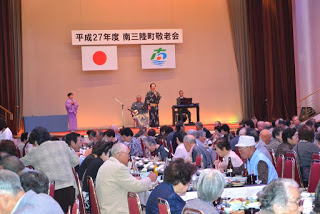 舞台の上で複数人が着物姿で歌や演奏をしており、客席では食事中の参加者がステージに注目している写真