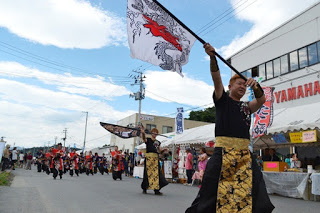 金と黒の衣装を着た旗振り隊の男性が大きな旗を高く掲げて歩き、後ろに踊り手の行列が続いている写真
