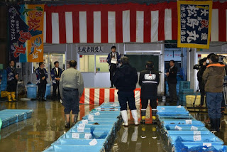紅白幕や大漁旗が見える屋内の魚市場で、中央の台の上に立つ男性が挨拶を行っている様子が写されており、周囲には水の入った青い容器を囲む人々が話を聞いている初競りの様子の写真