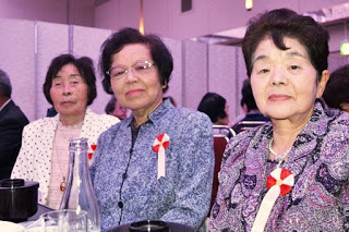 赤白のリボンを胸につけた三人の女性が整った髪型と上品な服装で食事テーブルに並んで座り、穏やかな表情でカメラを見つめている敬老会会場内の写真