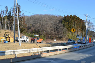 背景には木々や丘が広がり、道路沿いには曲がりくねった道を示すカーブの道路標識があり、ブルドーザーやトラックなどの建設車両や機材が並ぶ道路工事現場の様子の写真