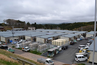 プレハブが並ぶ仮設住宅の敷地全体を高台から見下ろす構図で撮影しており、多数の軽自動車が仮設住宅横に停まっている復興住宅地の写真