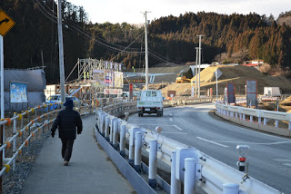 周囲には木々や丘が広がり、複数の看板や電柱が立っている道路沿いのガードレールのある歩道を歩く人物が写っており、道路には白い車が走っていて、背景には足場や機械が見える工事現場がある写真