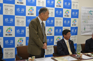 右側の男性が腕を前に組み立ちながら話をし、隣に男性が座っている様子を写した写真