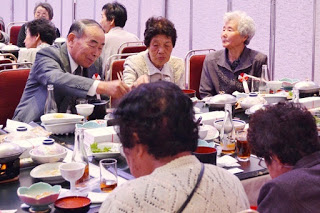 和やかな雰囲気の中で高齢者たちが料理を取り分け合いながら笑顔で食事を楽しんでいる様子の写真