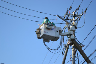 青空の下、安全装備を着用している二人の作業員がバケットリフトに乗って電柱に取り付けられた電線の作業をしている様子を写した写真