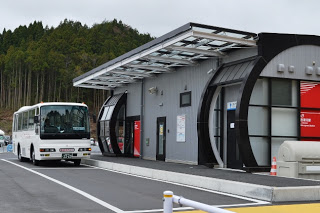 山のふもとの広い駐車スペースに停まっている白い中型バスと、黒とグレーを基調とした近代的なデザインの建物が写っているバス停留所の写真