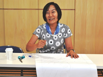 女性が室内のテーブルに座りながら笑顔で紙に下絵を描いており、周囲にはさみなど切り絵制作に使う道具が置かれている作業中の写真