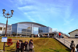 青空の下、全面ガラス張りの近代的な建物の前で数人の人々が立ち話をしていて、右側の階段には建物へ入る人の姿が見える写真