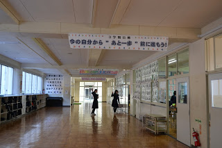 中学校の広い廊下の天井に「今の自分から"あと一歩"前に進もう！」と書かれた横断幕が掲げられており、奥にいる2人の女子生徒こちらに向かってピースサインをしている写真