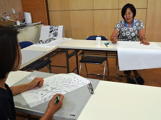 向かい合って座っている二人の女性が、それぞれ切り絵の作業をしながら会話を楽しんでいる様子の写真