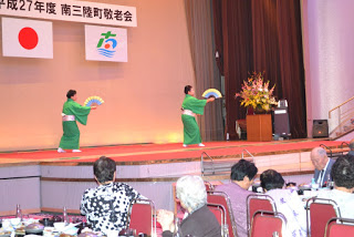 緑色の和服姿の女性2人がステージで扇子を広げて優雅に踊っており、会場の高齢者たちが食事をしながら観賞している敬老会の写真