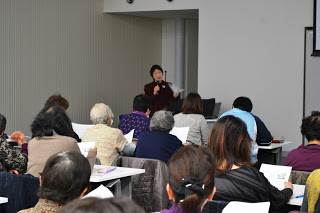 マイクを持って前に立った足立先生が、席に座ったたくさんの受講者たちに話をしている写真