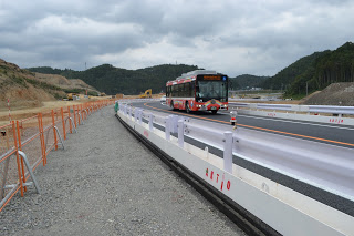 曇り空の下、赤と白のバスが両側にバリケードが設置された新しい道路を走っており、周囲にはオレンジ色の安全フェンスや建設機材が見られ、遠くには木々に覆われた丘が広がる工事現場付近の写真