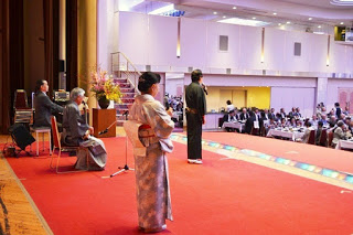 舞台の袖から着物姿の司会者と進行役が会場に向かって立ち、来賓がステージ横の椅子に座っている様子を収めた写真