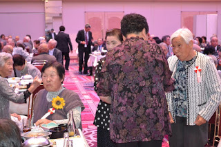 会場の一角で数人の高齢女性が立って談笑しており、周囲には座って食事を楽しむ参加者たちがいる敬老会の写真