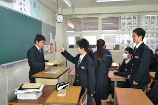 教室で制服を着た生徒たちが教壇に立った男子生徒が手に持つ黄色い募金箱に順番で募金をしている写真