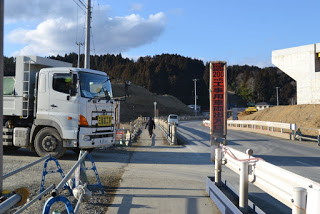 周囲は木々や丘に囲まれ、右側には建設機材が見え、赤い標識には「工事用車両出入口」と書かれている道路の左側にトラックが停まっており、道路を歩く人物と遠くにもう一台の車両が見える工事現場の様子の写真