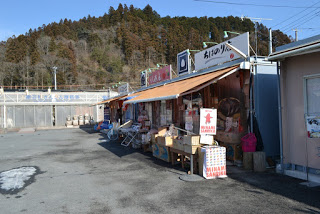 小高い山を背景に、屋根の上に看板を掲げ、軒下に商品を並べた店舗が複数並んでいる商店街の写真