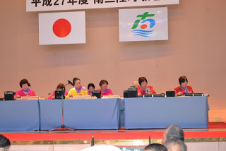 舞台上でピンク色と黄色のTシャツを着た複数の女性がテーブルに座り、南三陸大正琴を演奏している写真