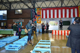 天井やステージに紅白幕や大漁旗が掲げられ、魚介類が入っている青い容器が床に並べられた、屋内の魚市場の初競り会場で、ステージに立つ人物が人々に向かって話しているおり、話を聞いている人たちが拍手を送っている様子の写真