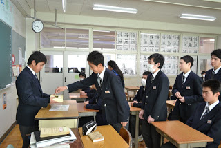 教室で制服を着た生徒たちが一列に並んで、教壇に立った男子生徒が手に持つ黄色い募金箱に順番で募金をしている写真