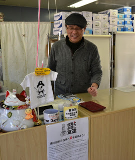 男性が立っている土産店のカウンターの猫の置物や小さいTシャツなどの横に募金箱が置かれ、その下に募金についての案内が貼ってある写真