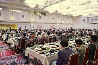 テーブルに座った多くの高齢者が料理を囲み、会話を楽しみながら賑やかに食事をしている全体の様子を捉えた写真