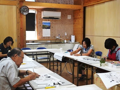 数名の男女が教室内の口型に並んだテーブルに向かい、それぞれ黙々と紙に下書きをしたりカッターで切ったりしながら切り絵制作に取り組んでいる教室の様子の写真