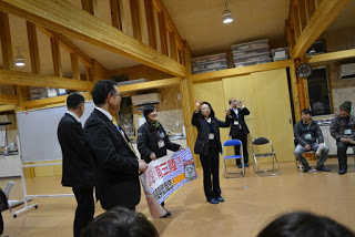 スーツを着た男性が2人の横に、横断幕を持った女性が立っており、その右側立った女性が、参加者たちが座る方向を向いて指揮を執るように両手を上げている様子の写真