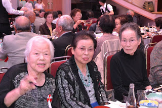 宴会場のテーブルで、正面を向いた高齢の女性3人が笑顔で座っている様子で、周囲では他の参加者が食事や会話をしている写真