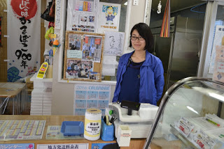 後ろの壁にたくさんの写真などが貼られた、かまぼこ店のカウンターに女性が立って立っており、カウンターのレジの横に募金箱が置かれている写真