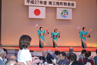 ステージ上で青緑色の和服を着た3人の女性が扇子を持って踊りを披露しており、観客が食事をしながら見守っている敬老会の写真