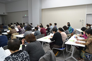 広い会場集まった大勢の参加者た、机に資料を広げて講義に臨んでいる様子の写真