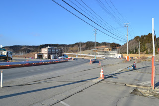 空は青く晴れ、背景には建物や木々のある丘が見え、道路に沿って電線と電柱が立ち並び、道路の脇に工事や補修作業を示すように三角コーンが並べられれている写真