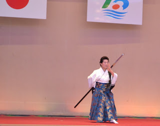 舞台の上で和装の女性が一人で正座し、脇に日本刀のような長い刀を置いて演舞をしている様子の写真