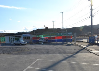 背景には電柱や電線、遠くに丘が見える敷地を囲むフェンスには赤・青・緑のカラフルなパネルや看板が取り付けられ、白い車が停まっている傍に小さな建物が建っている写真