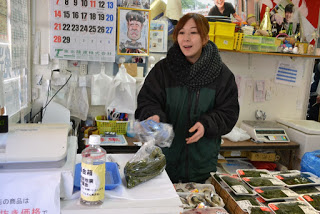 海藻が入った食品トレーがたくさん並んだ鮮魚店の店内で、女性が海藻の袋詰めを行っている白い台の上にペットボトルを利用した募金箱が置かれている写真