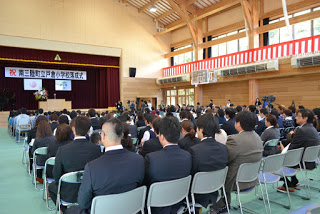木の梁が美しい広々とした体育館で、多くの来賓や保護者が整然と並び、壇上の演説を見守っている落成式会場内の写真