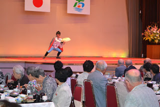 ステージの前で食事中の観客たちが見守る中、青と赤の衣装を着た人が元気よく踊っているパフォーマンスの写真