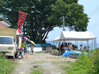 白いテントの下に人々が集まり、左側には「たいやき」と書かれた旗が立つ黄色い車が停まっている屋外の様子の写真